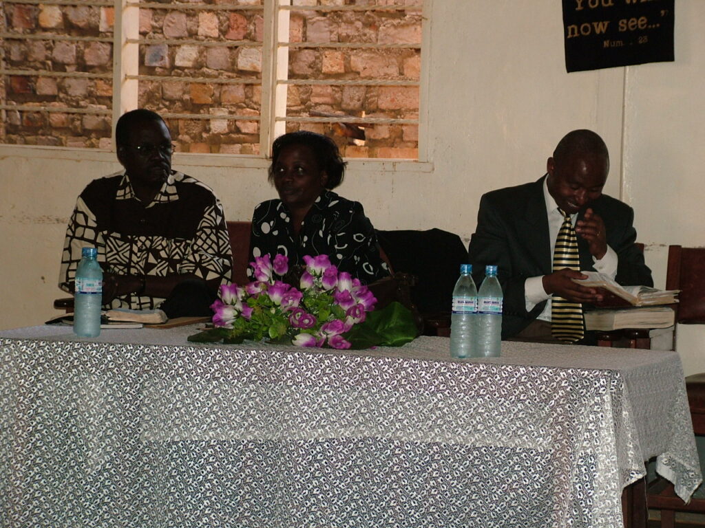 Pastor John Ekudu-Adoku seated in church with his wife Grace Ekudu.  John Habarurema on extreme right