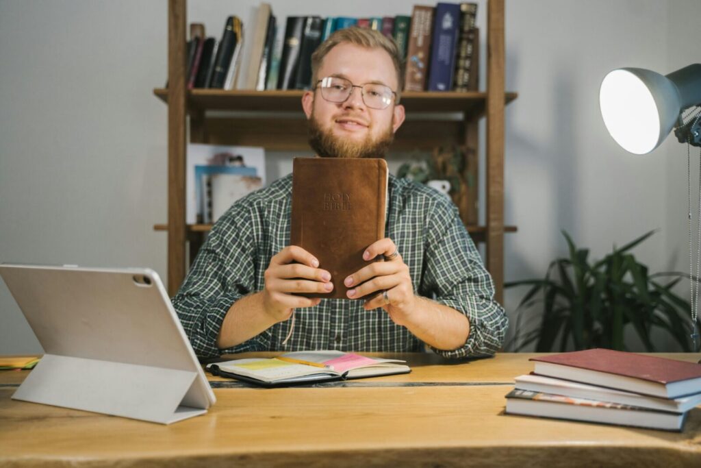 A man in eyeglasses holds a holy Bible at a wooden desk in a study room with shelves of books.