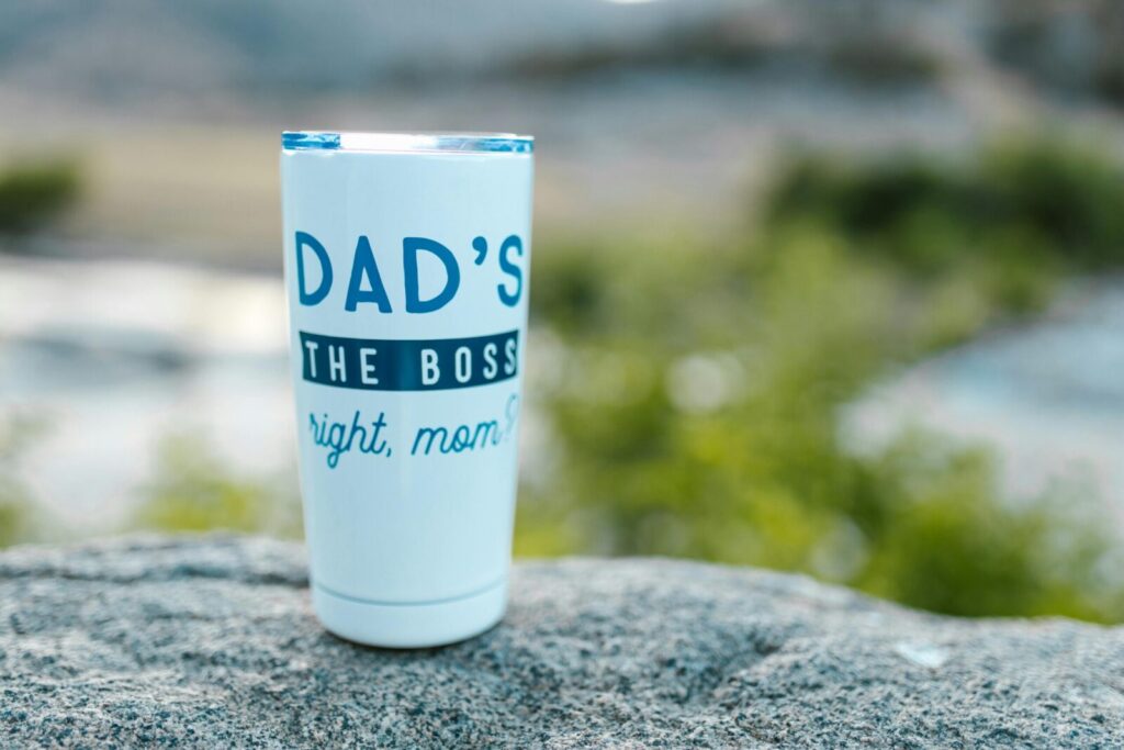 White tumbler with 'Dad's the Boss' text, set on rock in outdoor setting with greenery.
