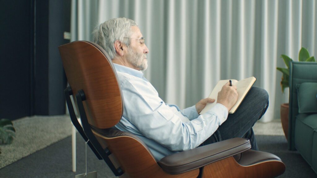 Senior man sitting in a modern chair writing in a notebook. Captured indoors with a calm atmosphere.